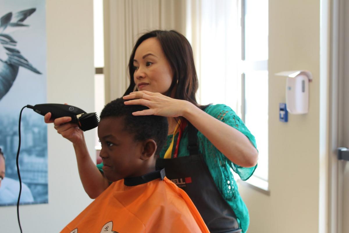 Haircut for RMHC Bay Area Child Smiling young boy gets a haircut from a stylist in a green blouse and apron inside a bright room.